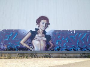 Ein Mädchen als kunstvolles Graffiti stützt die Arme in die Hüfte. Hintergrund türkis-graublaue Verzierung an kahler Betonwand